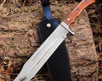 May include: A large, polished steel knife with a wooden handle and a black leather sheath. The knife has a wide blade and a full tang construction. The handle is a rich brown color with three small, blue-colored rivets. The sheath has a snap closure.