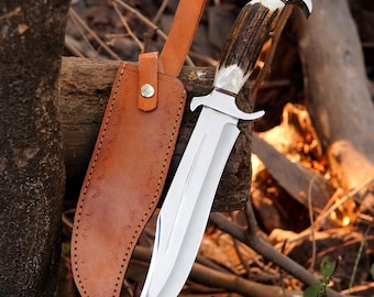 May include: A large hunting knife with a polished steel blade and a handle made of antler and bone. The knife is accompanied by a brown leather sheath with decorative stitching and a snap closure. The knife is approximately 12 inches long.
