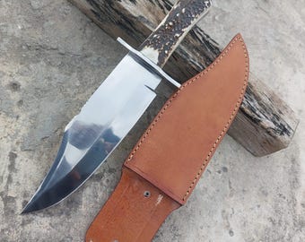 May include: A large, polished steel knife with a textured antler handle and a brown leather sheath. The knife has a wide blade and a sharp point. The sheath is stitched and has two rivets. The knife is resting on a piece of wood.