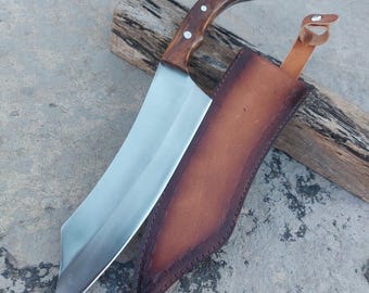 May include: A large, stainless steel cleaver with a curved, wooden handle and a brown leather sheath. The handle is secured with rivets. The sheath has a decorative stitched border and a leather loop.