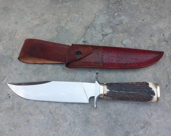 May include: A hunting knife with a brown leather sheath and a stag horn handle. The blade is silver and has a slight curve.