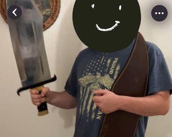 May include: A large, silver-colored metal knife with a wide blade and a black handle with gold-colored accents. The knife is accompanied by a long, brown leather sheath with visible stitching along the edges and numerical text printed on the surface.