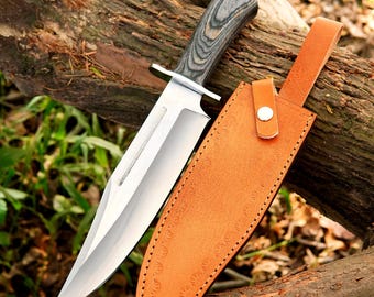 May include: A large hunting knife with a silver blade and a wooden handle with gray streaks. The knife is paired with a brown leather sheath with a snap closure. The knife is resting on a log.