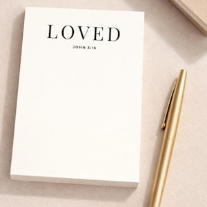 Könnte beinhalten: Ein weißer Notizblock mit dem Wort "LOVED" in Schwarz gedruckt, zusammen mit "JOHN 3:16". Ein goldener Stift liegt neben dem Notizblock. Der Notizblock und der Stift befinden sich auf einer hellen Oberfläche.