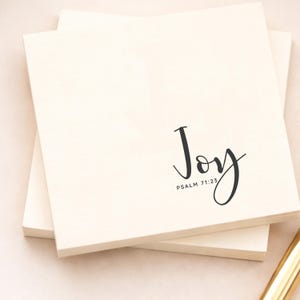 May include: Cream-coloured notepads stacked on top of each other. The word "Joy" is written in a stylish, black script, with "Psalm 71:23" below it. The notepads are on a light-coloured surface.
