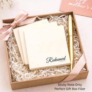 Puede incluir: Una caja de regalo con exterior marrón claro, llena de papel triturado, una pila de notas adhesivas color crema con la palabra "Redeemed" impresa y un bolígrafo dorado. Una cinta y un lazo rosas adornan la caja.