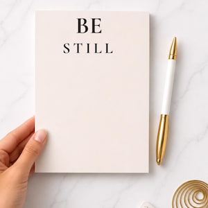 May include: A notepad with the words "BE STILL" in black font. A white and gold pen rests beside the notepad. The notepad is a light peach color. The background is a white marble surface.