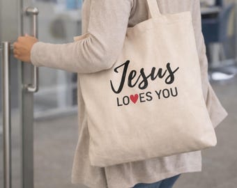 Może przedstawiać: Beżowa torba typu tote z napisem "Jesus LOVES YOU" w czarnym skrypcie i czerwonym sercem. Torba ma prosty design i jest noszona na ramieniu.