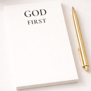 May include: A white notepad with the words "GOD FIRST" printed in black. A gold-colored pen rests beside the notepad. The notepad and pen are on a white surface.