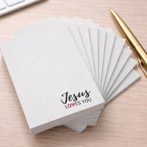 May include: A stack of white notepads with the text "Jesus loves you" in black script and a red heart. A gold pen rests on a light wood surface next to the notepads. The notepads are blank and ready for use.