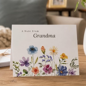 May include: A folded greeting card with the text "A Note From Grandma" above a watercolor floral design. The flowers include blue, purple, orange, and yellow blooms. The card is on a wooden surface.