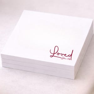 Könnte beinhalten: Ein Stapel weißer quadratischer Notizblöcke. Das Wort "Loved" ist in kursiver, burgunderroter Schrift auf dem obersten Notizblock geschrieben, darunter "John 3:16". Die Notizblöcke sind auf einer weißen Oberfläche gestapelt.