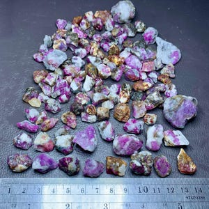 Puede incluir: Una colección de piedras preciosas en bruto, sin tallar, en tonos rosa, morado, blanco y marrón. Las piedras varían en tamaño y forma, con una regla de acero inoxidable para la escala. Las piedras preciosas están dispersas sobre una superficie oscura.