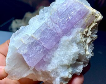 May include: A raw, natural mineral specimen featuring a central section of lavender-colored crystals embedded in a matrix of white and beige rock. The crystals have a rectangular shape and a layered appearance.