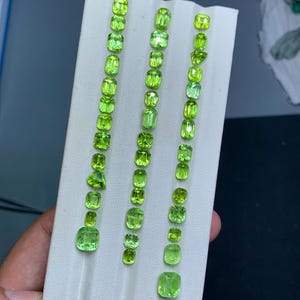 Puede incluir: Filas de gemas verde lima vibrantes de varias formas y tamaños se exhiben sobre una superficie blanca. Las gemas están dispuestas en líneas verticales, mostrando su claridad y color. Las gemas son probablemente para la fabricación de joyas.