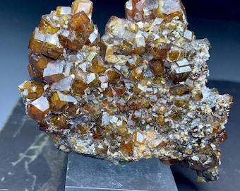 May include: A cluster of lustrous, amber-colored crystals, likely a mineral specimen, with a complex, multi-faceted structure. The crystals are set against a matrix of darker, earthy tones, creating a striking contrast. The specimen is displayed on a gray rectangular base.