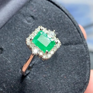 May include: A square emerald gemstone ring with a halo of smaller diamonds set in white gold.