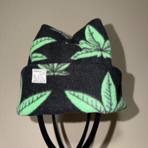 May include: A black beanie with a repeating light green leaf pattern. The hat features a white rectangular patch on the front cuff with a black line drawing of a mountain, a pine tree, and a small orange circle, with the text WanderLid above.