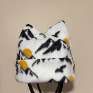 May include: A white fleece beanie featuring a mountain and sunset design in black and yellow. The hat has a folded brim and a small tag on the side. The design includes black mountain silhouettes, yellow suns, and flying birds.