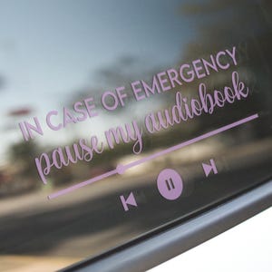 May include: A light purple car window decal with the text "IN CASE OF EMERGENCY pause my audiobook" in a cursive font. Below the text is a graphic of a media player with play, pause, and skip buttons.