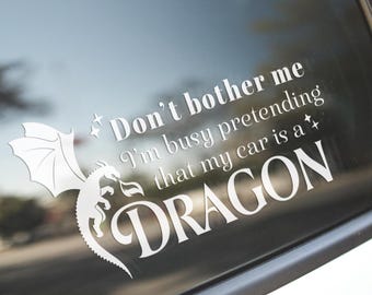 May include: A white vinyl decal applied to a car window featuring a silhouette of a dragon with wings spread and the text: Don't bother me I'm busy pretending that my car is a DRAGON, accented by small four-pointed star shapes on both sides.