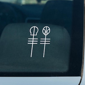 May include: A white vinyl decal sticker applied to a dark glass surface. The design features two vertical line symbols, each topped with a distinct geometric shape. The left symbol has a rounded top, while the right symbol has an oval top.