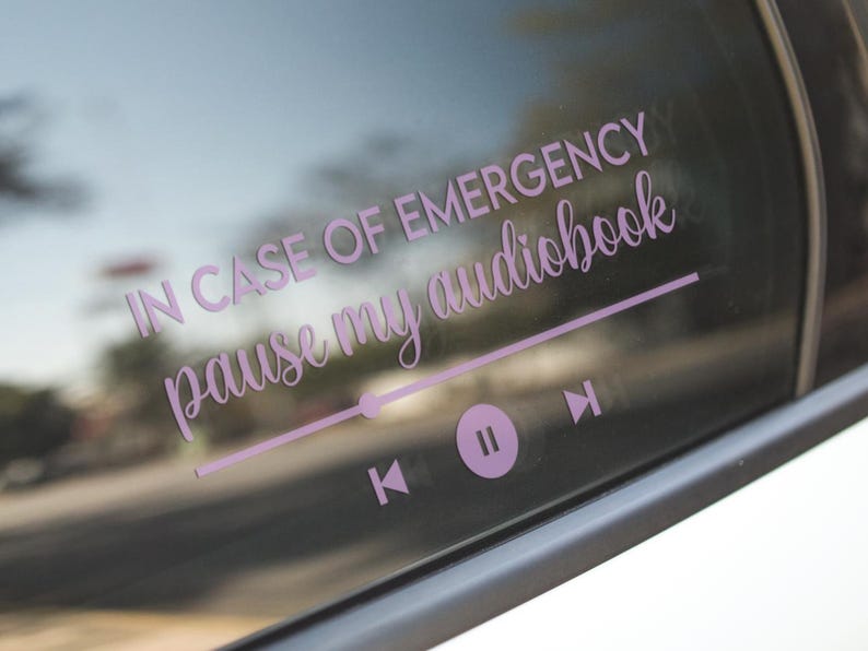 May include: A light purple car window decal with the text "IN CASE OF EMERGENCY pause my audiobook" in a cursive font. Below the text is a graphic of a media player with play, pause, and skip buttons.