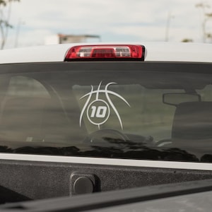 May include: A white vinyl decal applied to the rear window of a white pickup truck. The decal features a stylized basketball graphic with the number 10 centered inside a circular frame, rendered in clean, bold white lines against the glass.