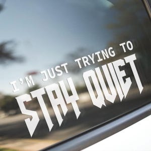 May include: White vinyl decal with the text "I'M JUST TRYING TO STAY QUIET" in a bold, stylized font. The decal is applied to a car window, with the background showing a blurred outdoor scene.