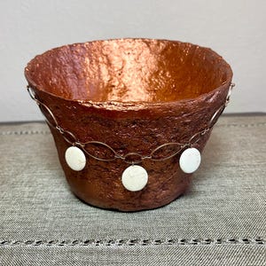 May include: A handcrafted, textured copper-colored bowl with a decorative chain featuring white, circular accents. The interior has a metallic sheen. The bowl is designed for decorative purposes.