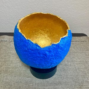 May include: A handcrafted, spherical bowl with a vibrant blue exterior and a shimmering gold interior. The rim has a textured, uneven edge. The bowl is mounted on a small, black base, creating a striking contrast of colors and textures.