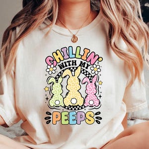 May include: A white t-shirt with the text "Chillin' with my peeps" in rainbow colors and a graphic of three pastel colored bunnies with black polka dots. The bunnies are standing on a black and white checkered pattern.