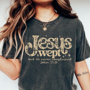 May include: Dark gray t-shirt with the text "Jesus wept" in a decorative font, with floral accents. The phrase "but He never complained John 11:35" is printed below. The shirt is paired with blue jeans and white shoes.