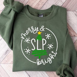 May include: A green sweatshirt with the words "Merry & SLP bright" in white script and a Christmas tree graphic. The design is inside a white circle with snowflakes. The sweatshirt has cuffed sleeves and a crew neck.
