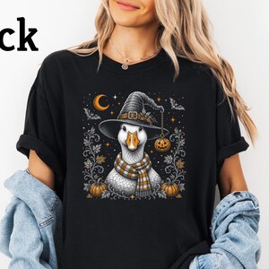 Puede incluir: Una camiseta negra con un estampado gráfico de un ganso blanco que lleva un sombrero de bruja y una bufanda de cuadros. El diseño incluye calabazas naranjas, una luna creciente, estrellas y murciélagos, con la palabra Black arriba.
