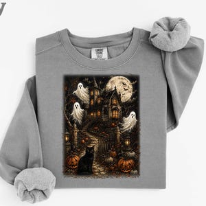 Puede incluir: Una sudadera gris de cuello redondo con un estampado gráfico de una casa encantada por la noche. La ilustración incluye una luna llena, tres fantasmas blancos, un gato negro sentado en un camino de piedra y calabazas naranjas entre árboles.