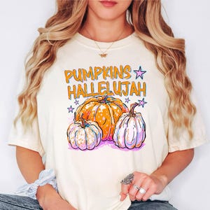 Puede incluir: Una camiseta de manga corta de color crema con un estampado gráfico que incluye el texto naranja PUMPKINS HALLELUJAH. El diseño presenta tres calabazas en tonos naranja, blanco y morado, acentuadas por pequeñas estrellas rosas y moradas.
