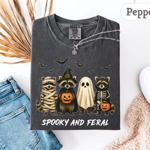 May include: Dark gray t-shirt with a Halloween-themed graphic. The design features four raccoons dressed as a mummy, a witch, a ghost, and a skeleton, holding pumpkins. The text "Spooky and Feral" is printed below the illustration.