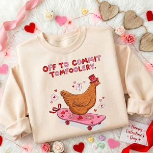 May include: A cream-colored sweatshirt with the text "OFF TO COMMIT TOMFOOLERY." features a cartoon chicken wearing a hat and riding a pink skateboard decorated with hearts. The design includes hearts and Valentine's Day elements.