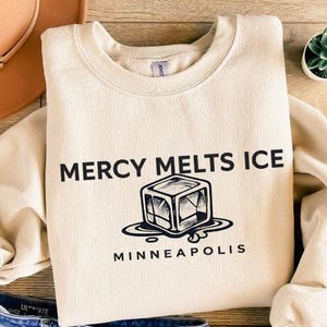 May include: Cream-colored sweatshirt with the text "MERCY MELTS ICE" and a graphic of a melting ice cube, with "MINNEAPOLIS" below. The sweatshirt is paired with blue jeans, a brown hat, and sandals.