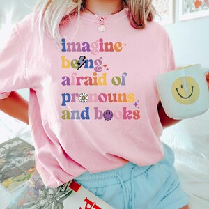 May include: A pale pink t-shirt featuring the phrase "imagine being afraid of pronouns and books" in a multi-coloured, retro style. The shirt is worn with light blue shorts and a white mug with a yellow smiley face.