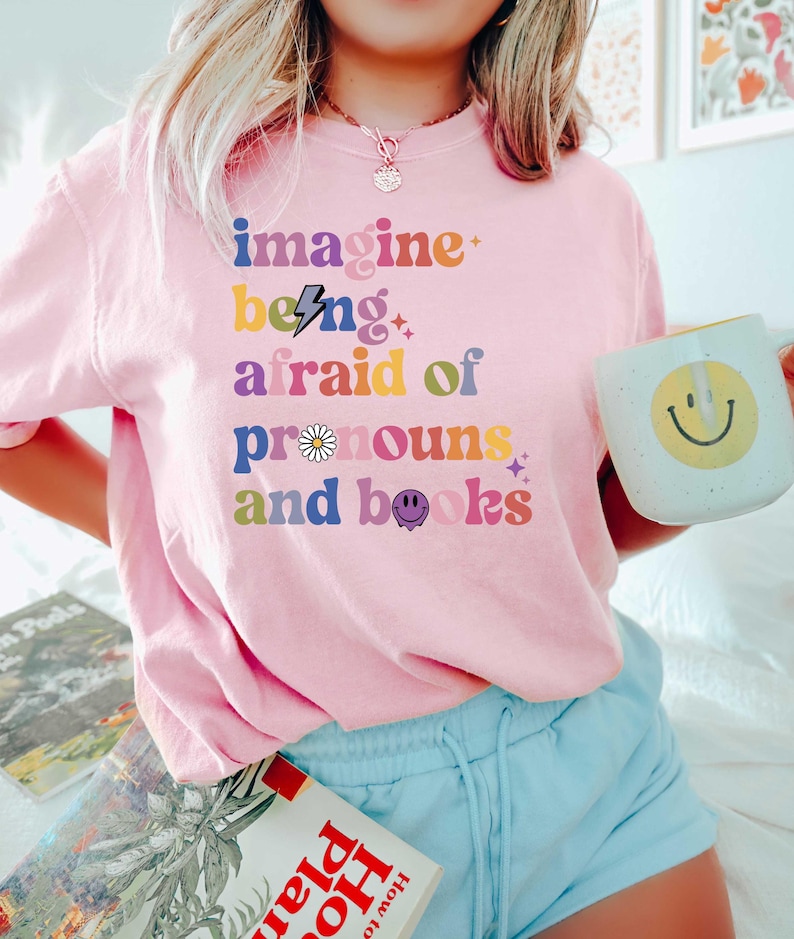 May include: A pale pink t-shirt featuring the phrase "imagine being afraid of pronouns and books" in a multi-coloured, retro style. The shirt is worn with light blue shorts and a white mug with a yellow smiley face.