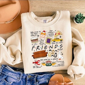 May include: Cream-colored Friends-themed sweatshirt with various cartoon illustrations and text. The design includes a sofa, pizza, and the Central Perk logo. The text includes phrases like "Welcome to the Real World!" and "How you doin?"