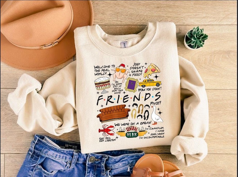 May include: Cream-colored Friends-themed sweatshirt with various cartoon illustrations and text. The design includes a sofa, pizza, and the Central Perk logo. The text includes phrases like "Welcome to the Real World!" and "How you doin?"