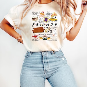 May include: Cream-colored t-shirt featuring a graphic design with the word "FRIENDS" and various illustrations from the TV show, including a couch, lobster, and Central Perk logo. The shirt also has several quotes from the show.