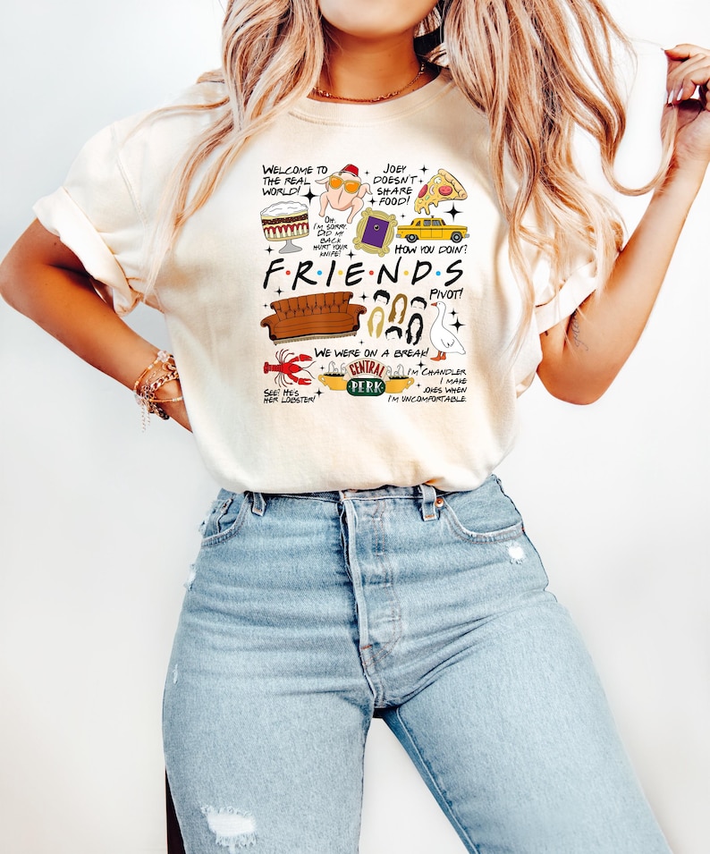 May include: Cream-colored t-shirt featuring a graphic design with the word "FRIENDS" and various illustrations from the TV show, including a couch, lobster, and Central Perk logo. The shirt also has several quotes from the show.