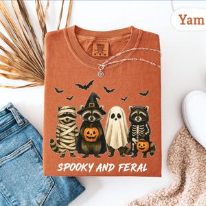 May include: A yam-colored t-shirt featuring a Halloween-themed graphic. The design includes four cartoon raccoons dressed as a mummy, witch, ghost, and skeleton, with bats flying above. The text "Spooky and Feral" is printed below the characters.
