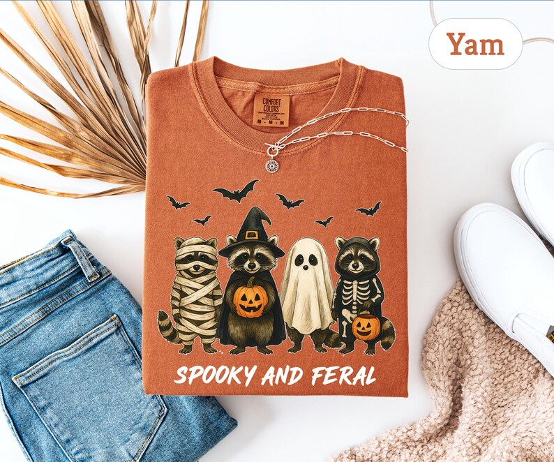 May include: A yam-colored t-shirt featuring a Halloween-themed graphic. The design includes four cartoon raccoons dressed as a mummy, witch, ghost, and skeleton, with bats flying above. The text "Spooky and Feral" is printed below the characters.