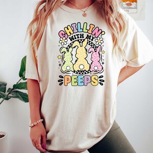 May include: A white t-shirt with a graphic of three pastel-colored bunnies with the text "Chillin' with my peeps" in rainbow colors.