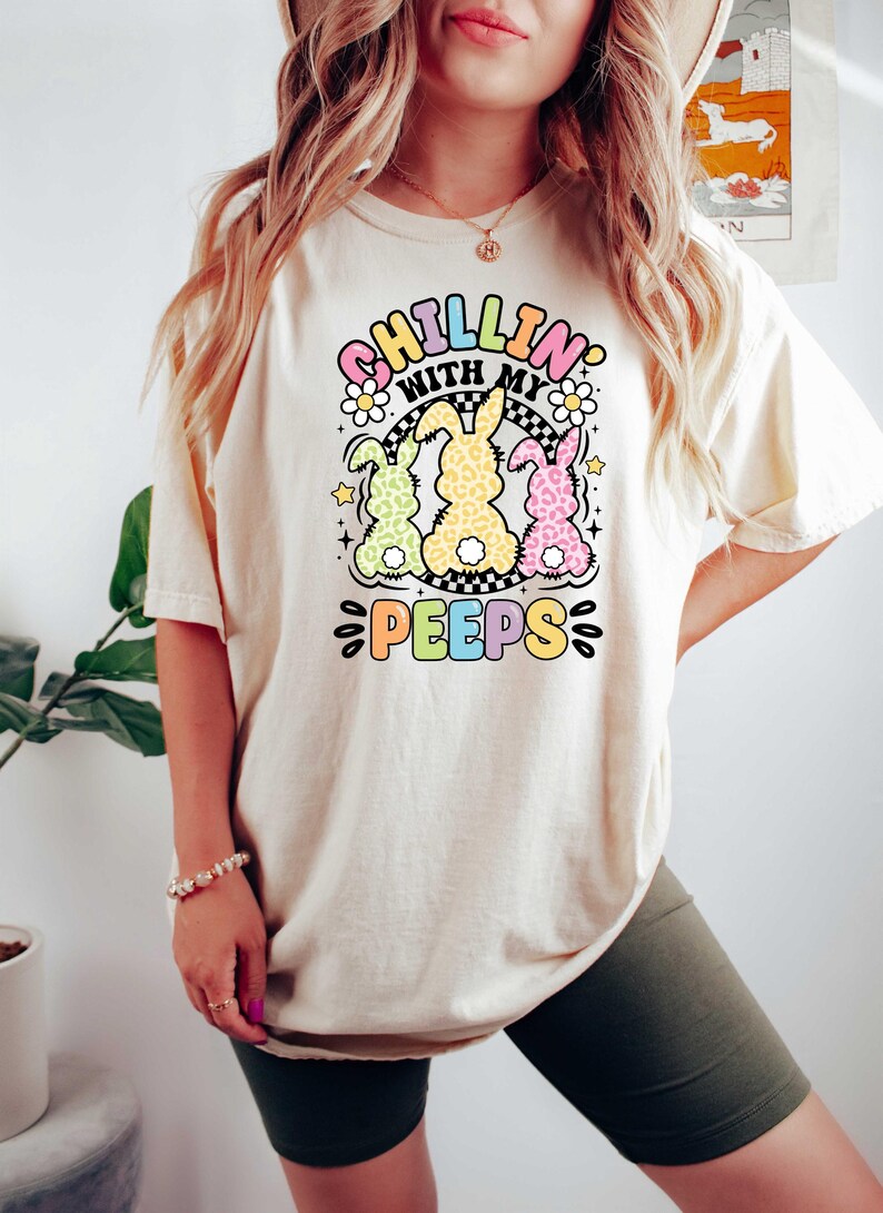 May include: A white t-shirt with a graphic of three pastel-colored bunnies with the text "Chillin' with my peeps" in rainbow colors.